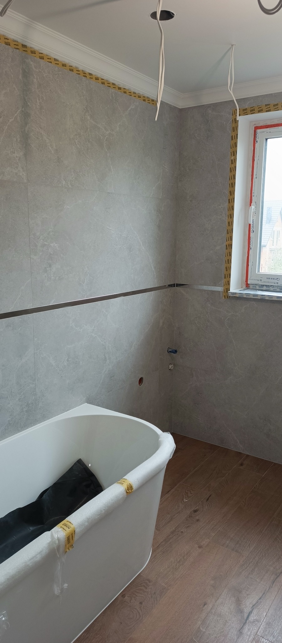 Szare płytki imitujące marmur na ścianach łazienki w trakcie remontu. Widoczna wanna, fragment okna i drewnopodobna podłoga. Taśma malarska zabezpiecza sufit.