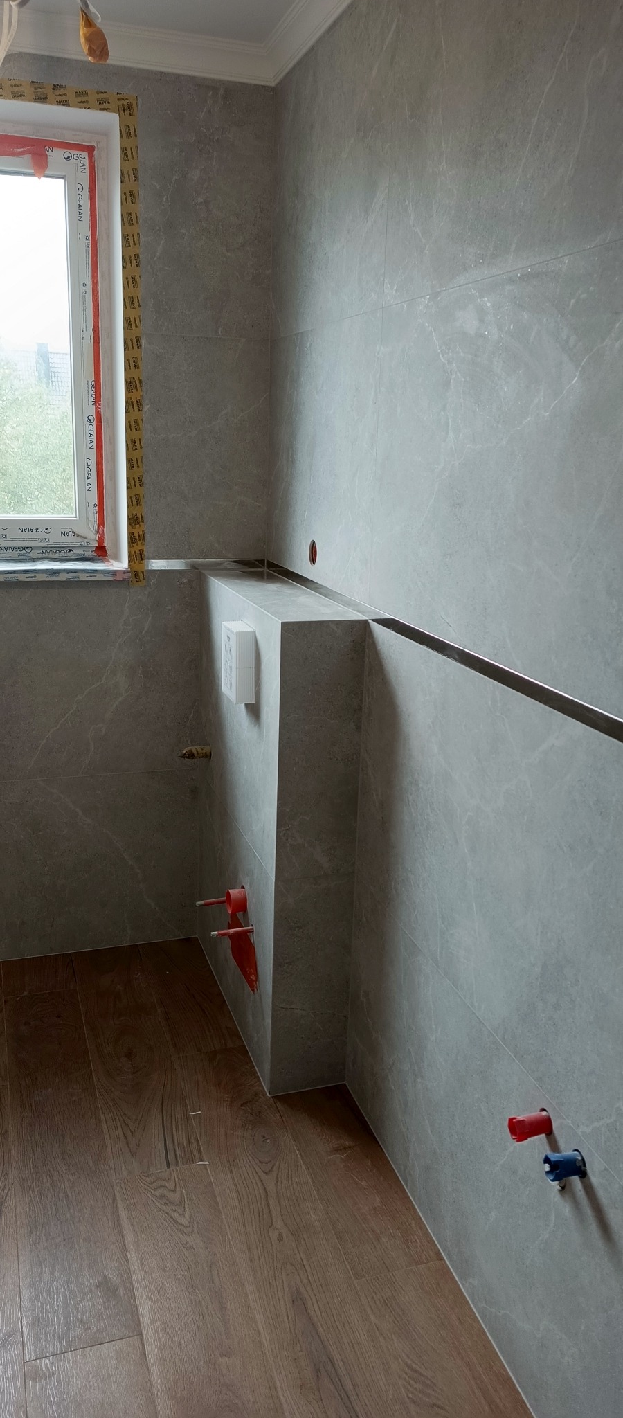 Wykończona łazienka z szarymi płytkami imitującymi marmur, drewnopodobną podłogą i zabudową WC. Widoczne przyłącza wodne i okno z żółtą taśmą zabezpieczającą.