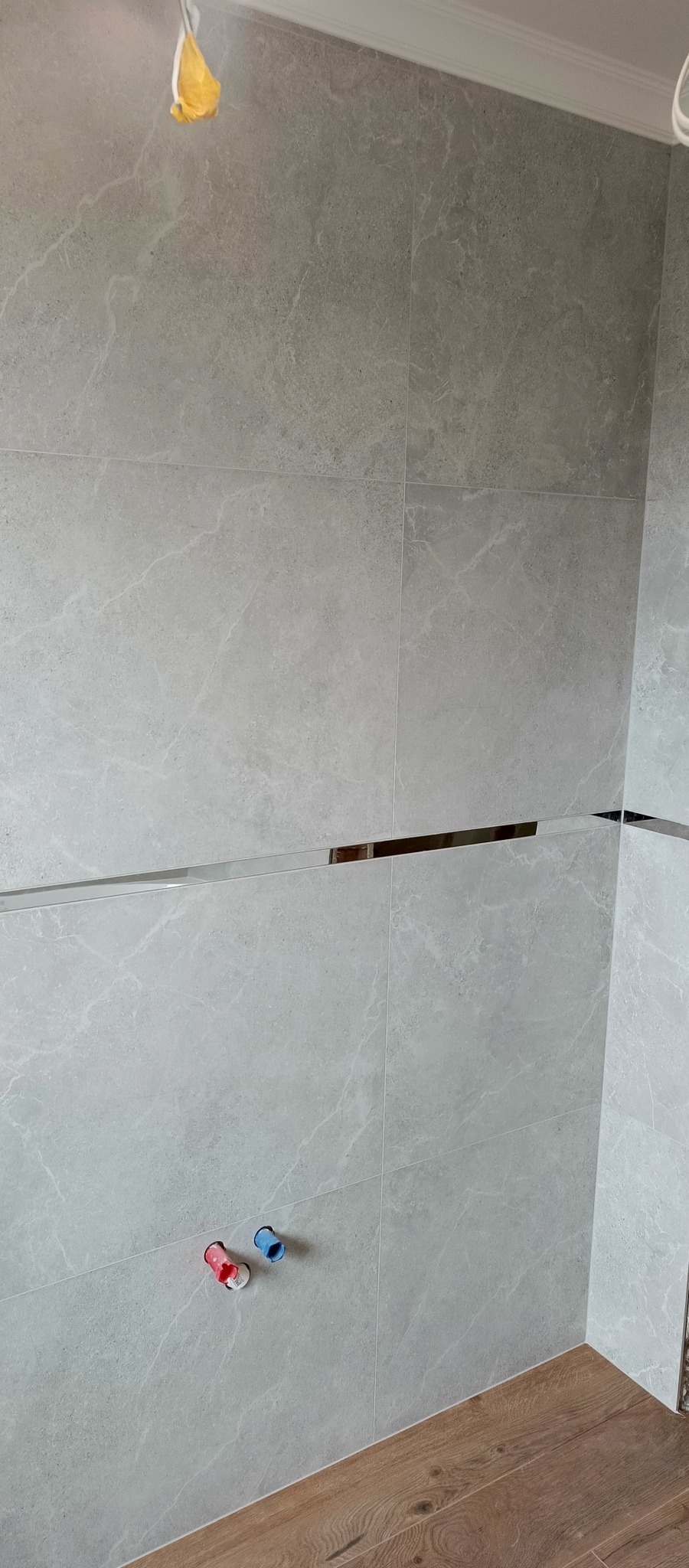 Ściana wyłożona szarymi płytkami imitującymi marmur, z dekoracyjną, poziomą listwą. Widoczne wystające rurki instalacji wodnej i fragment podłogi z paneli.