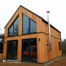 Nowoczesny dom szkieletowy z pionową elewacją z jasnego drewna, dużymi oknami z czarnymi ramami i kominem ze stali nierdzewnej, w tle widoczny pomarańczowy samochód.