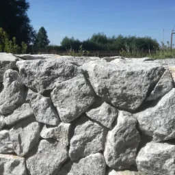 Niski mur oporowy z nieregularnych, szarych kamieni, z widoczną fakturą i spoinami, na tle zieleni i błękitnego nieba.