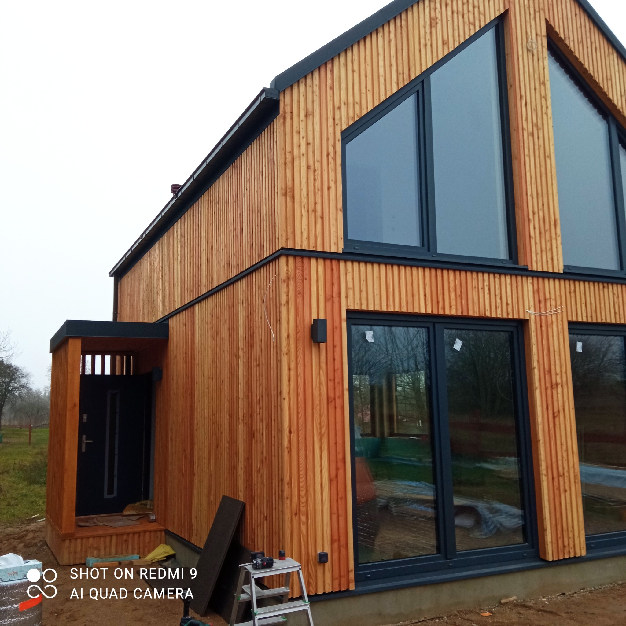 Nowoczesny dom z drewnianą elewacją w pionowe deski, z dużymi oknami w czarnych ramach i zadaszonym wejściem, w trakcie budowy, widoczne narzędzia i materiały budowlane.