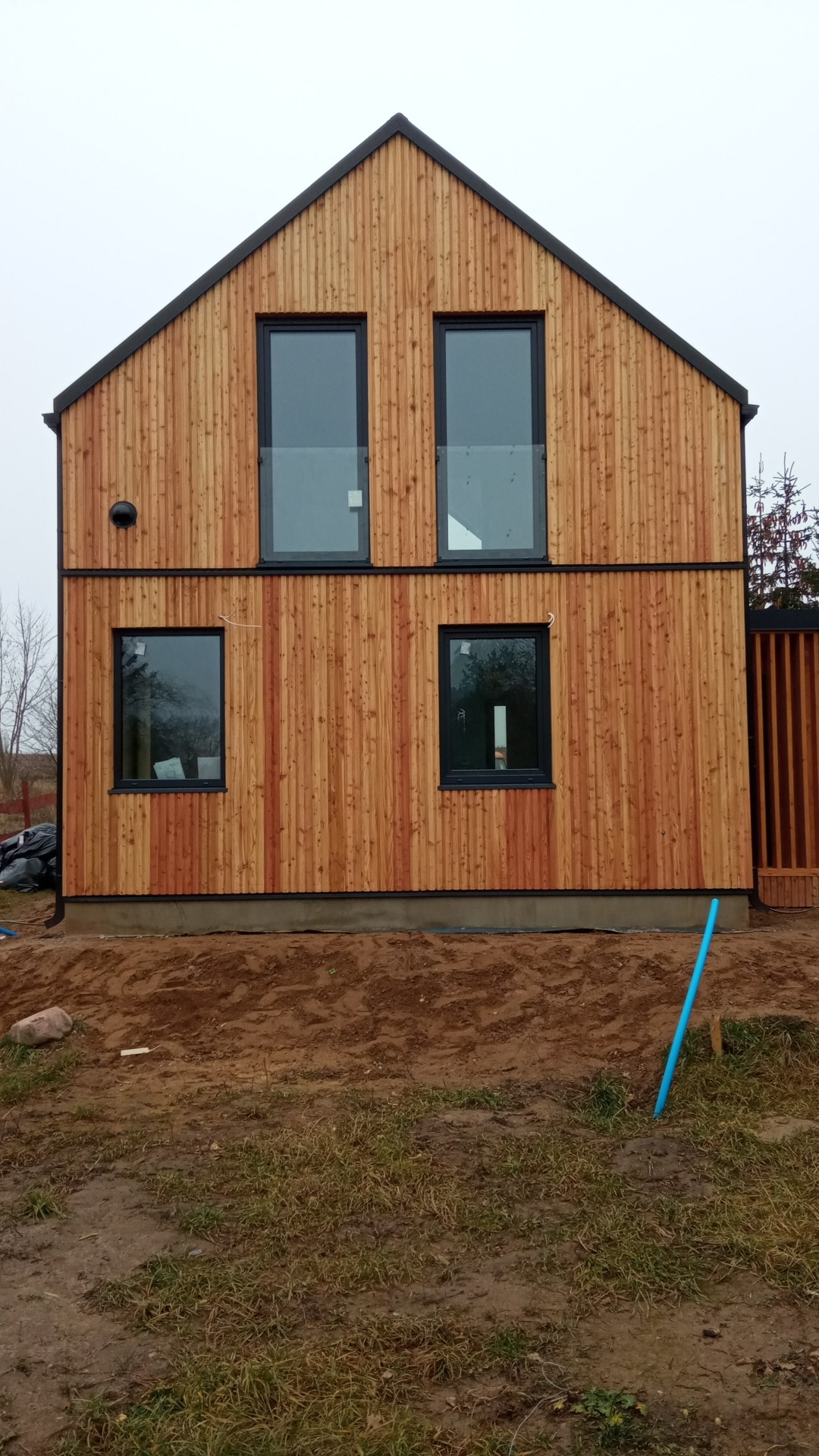 Dwukondygnacyjny budynek z drewnianą elewacją w pionowe deski, czarne obramowania okien i dachu, widoczny fragment fundamentu i teren wokół budynku.