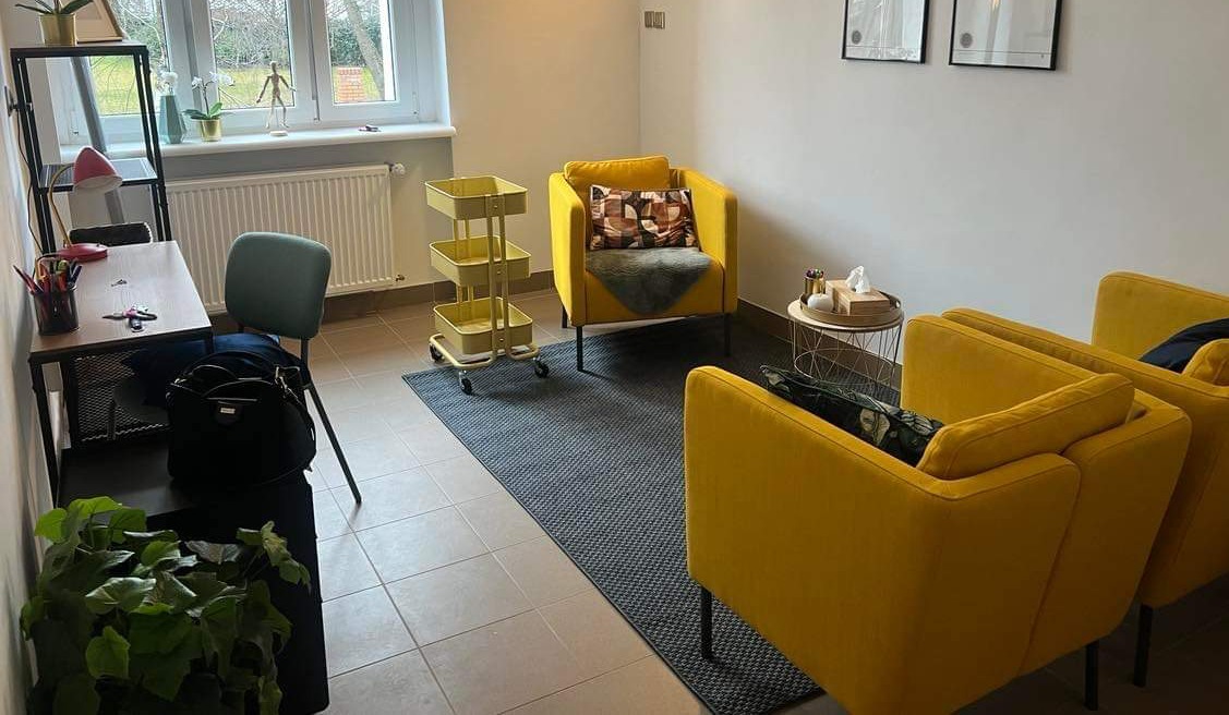 Jasne wnętrze gabinetu z żółtymi fotelami, biurkiem, regałem i wózkiem na kółkach, w tle okno z widokiem na drzewa.