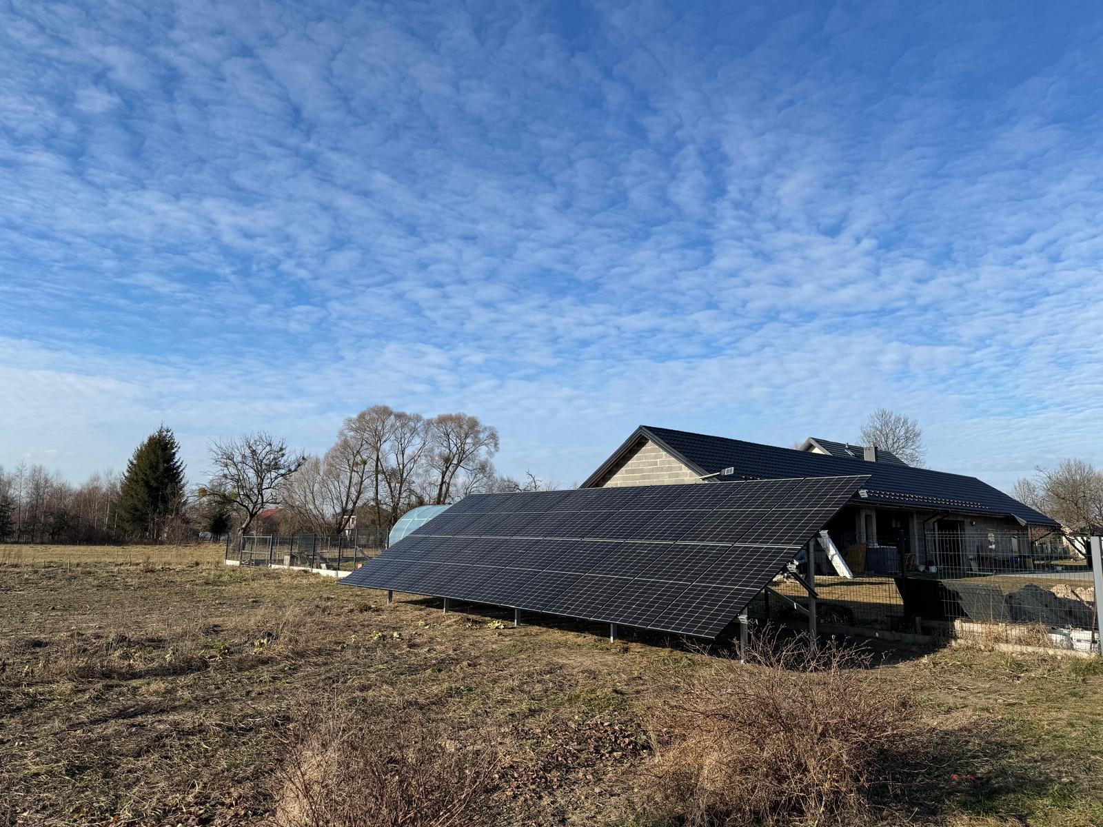Naziemna instalacja fotowoltaiczna z czarnymi panelami, zamontowana przed domem z szarej cegły, na tle pochmurnego nieba.