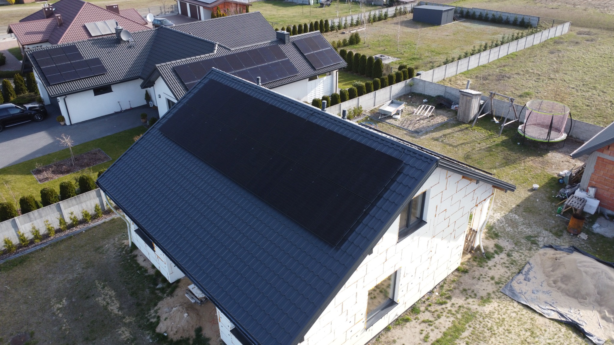 Widok z góry na dom z ciemnymi panelami słonecznymi na dachu z szarej dachówki. Dom w trakcie budowy, ocieplony styropianem. W tle inne domy i zielony ogród.