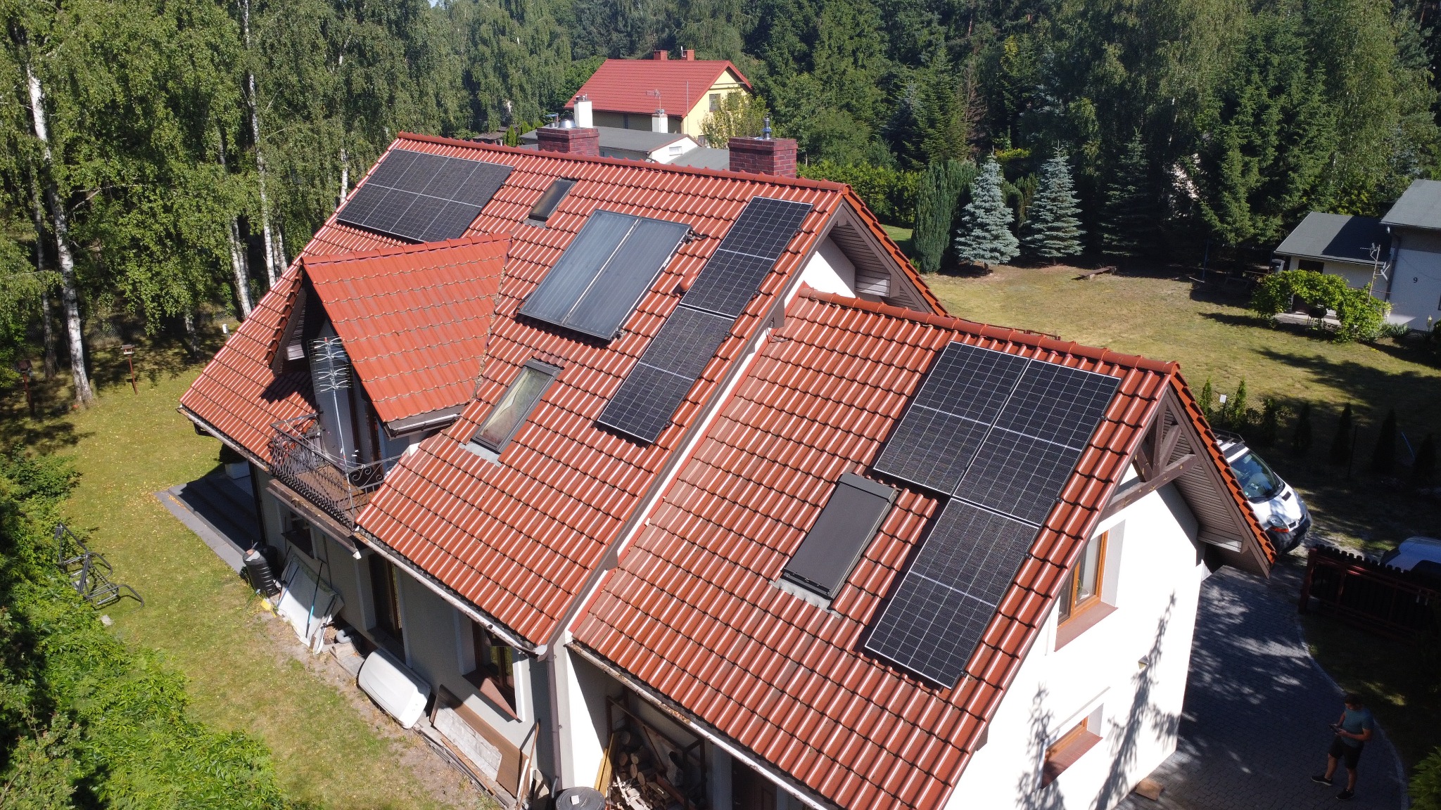 Czerwony dach domu z panelami słonecznymi i oknami dachowymi, widok z góry. Otoczenie: drzewa, trawnik, inne budynki. Różne typy paneli na dachu.