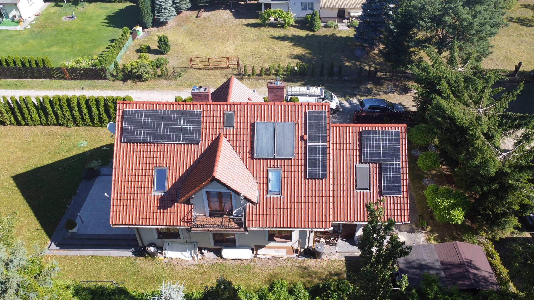 Widok z góry na dom z czerwoną dachówką i panelami słonecznymi. Różne rozmiary paneli, okna dachowe. Otoczenie: trawnik, drzewa, zaparkowane pojazdy.