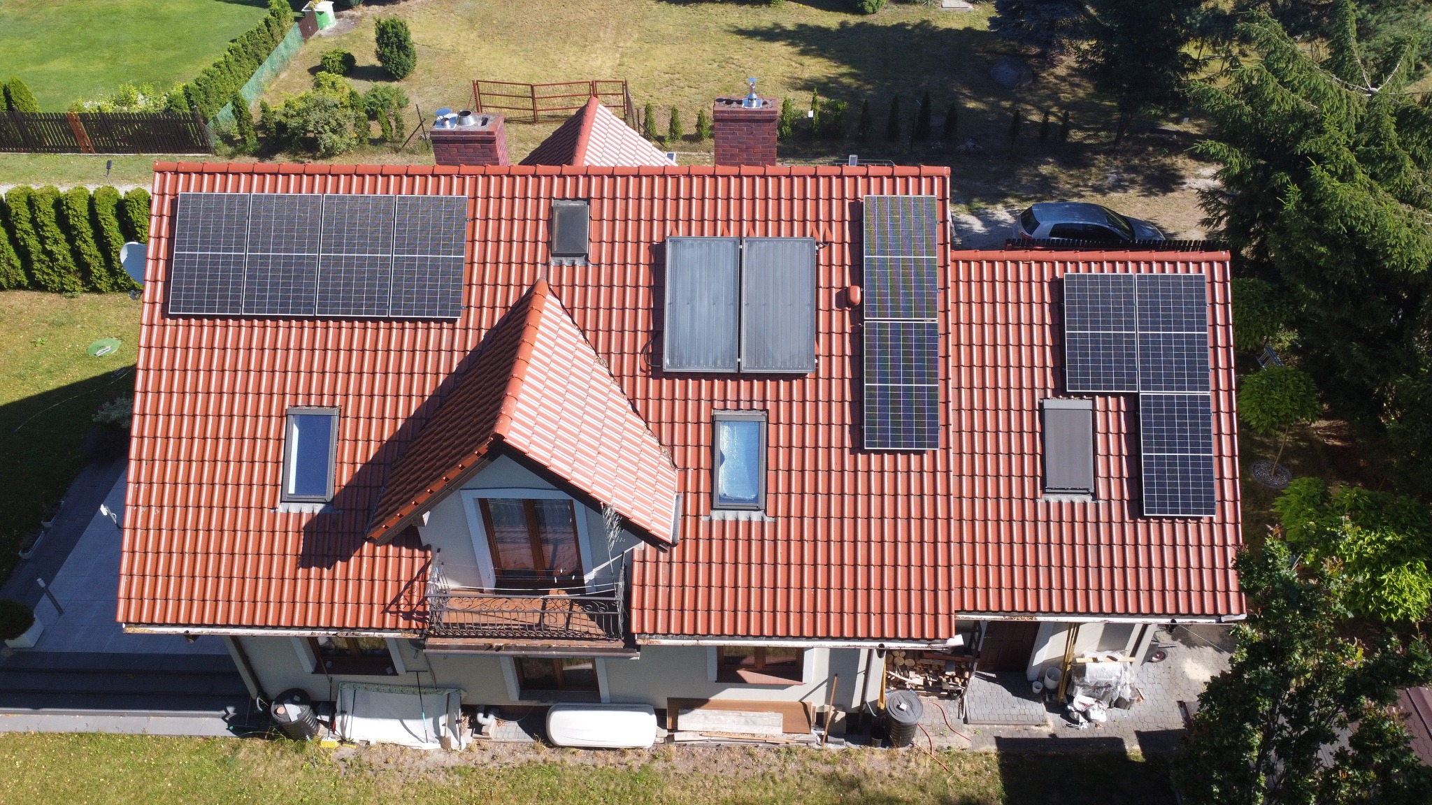 Czerwony dach domu z panelami słonecznymi i kolektorami, widok z góry. Różne rozmiary paneli, okna dachowe, kominy. W tle zieleń drzew i trawnik.