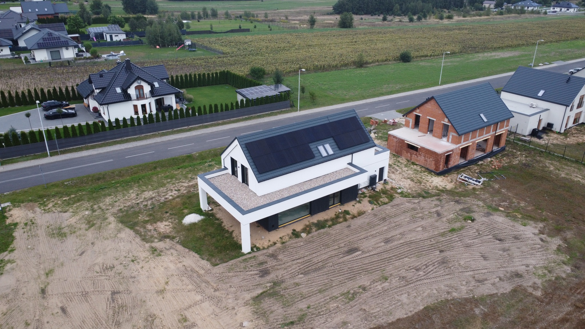 Nowoczesny dom jednorodzinny z panelami słonecznymi na dachu, widok z drona. W tle inne domy i pole uprawne. Minimalistyczna architektura, jasna elewacja.