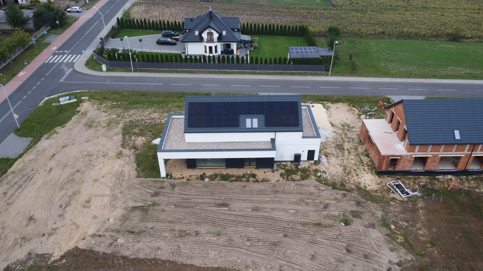 Widok z drona na nowoczesny dom z panelami słonecznymi na dachu w Bełchatowie. Obok budynek w stanie surowym. Widoczna droga i zabudowa jednorodzinna.