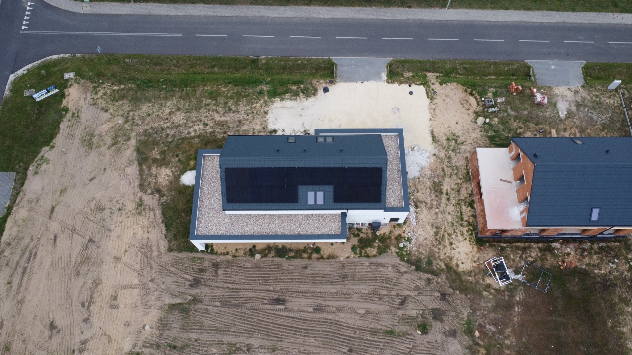 Widok z góry na nowoczesny dom z panelami słonecznymi na dachu, obok drugi budynek w trakcie budowy. Teren wokół w trakcie zagospodarowania, widoczna droga.
