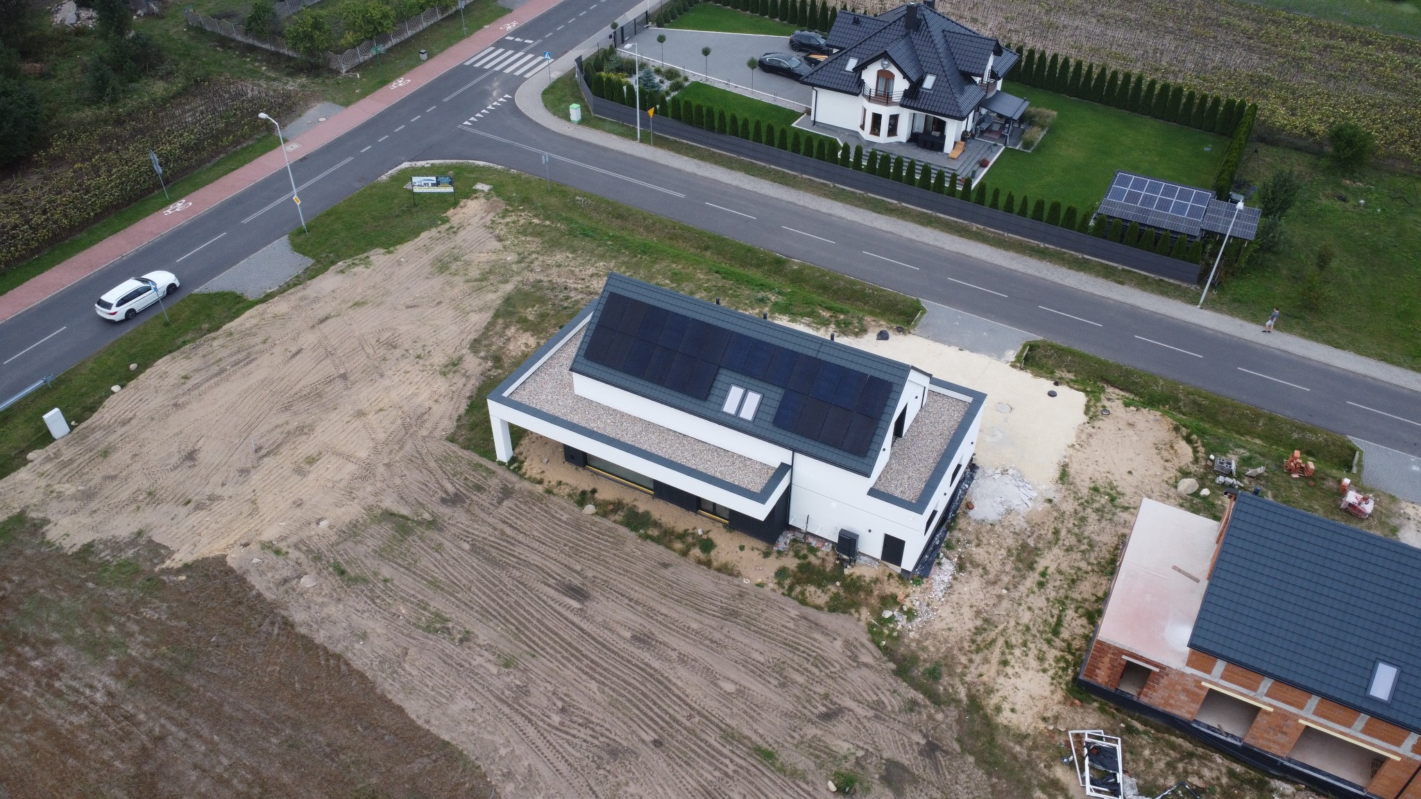 Nowoczesny dom z panelami fotowoltaicznymi na płaskim dachu, widok z drona. W tle drugi dom z panelami na gruncie i droga z jadącym białym samochodem.
