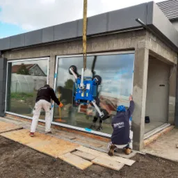 Montaż dużego, panoramicznego okna z użyciem podnośnika próżniowego; dwóch monterów zabezpiecza ramę, widoczne płyty OSB zabezpieczające podłoże.