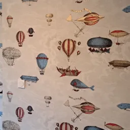 Ściana pokryta tapetą w jasnych odcieniach z motywem retro balonów, sterowców i latających ryb, widoczny fragment listwy przypodłogowej i gniazdka elektrycznego.