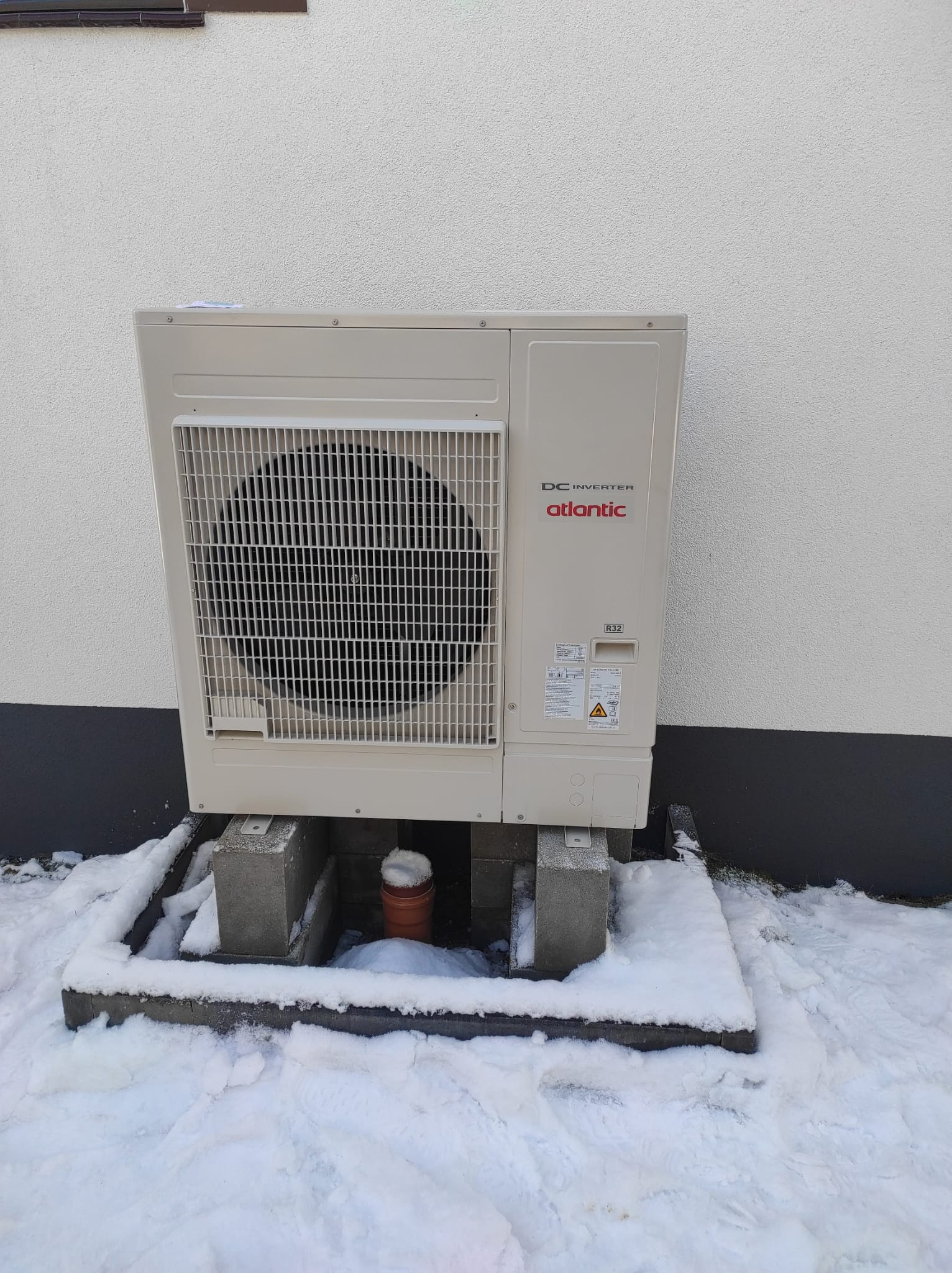 Zewnętrzna jednostka klimatyzacji Atlantic DC Inverter na betonowych podstawach, otoczona śniegiem przy ścianie budynku.