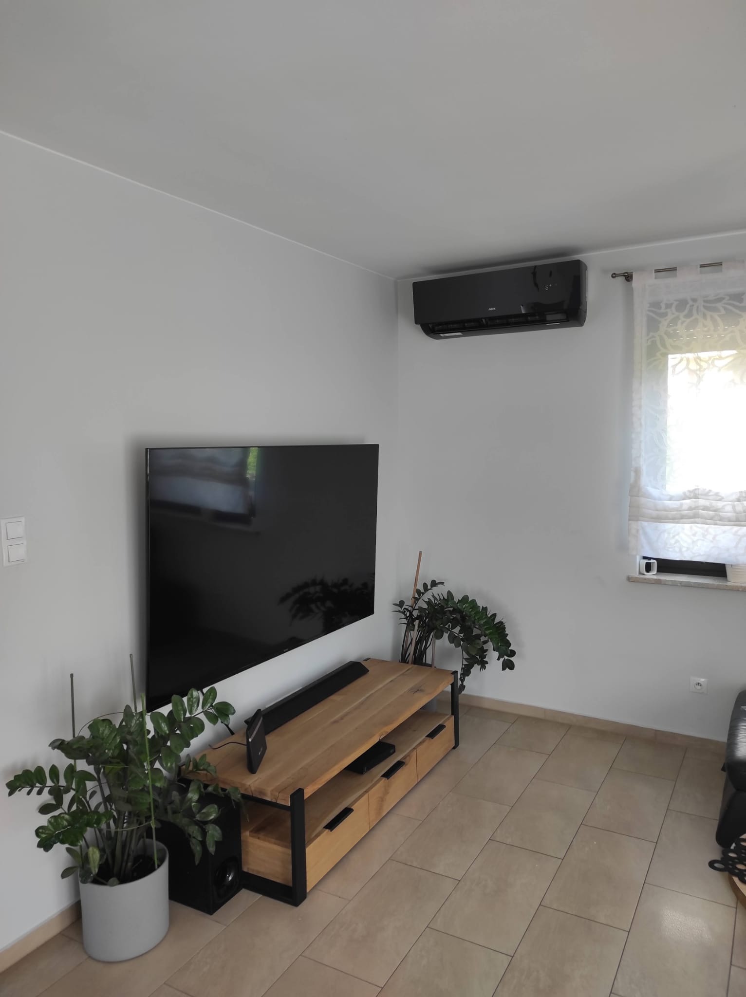 Czarny klimatyzator ścienny zamontowany w nowoczesnym salonie z telewizorem i drewnianą szafką RTV, widoczne rośliny doniczkowe.