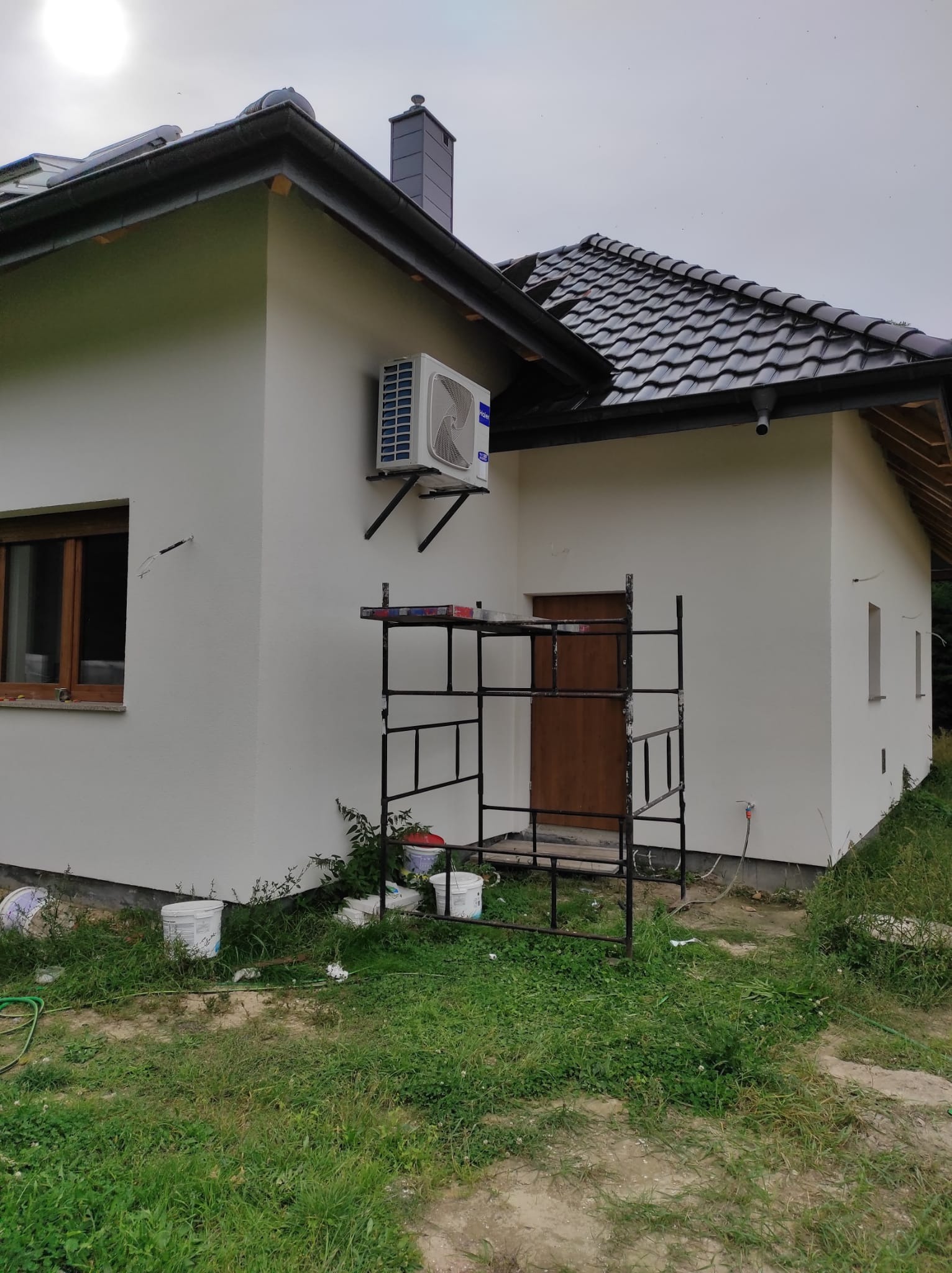 Biały dom jednorodzinny z zamontowaną klimatyzacją na ścianie, widoczne rusztowanie, kubełki po farbie i świeżo pomalowana elewacja.