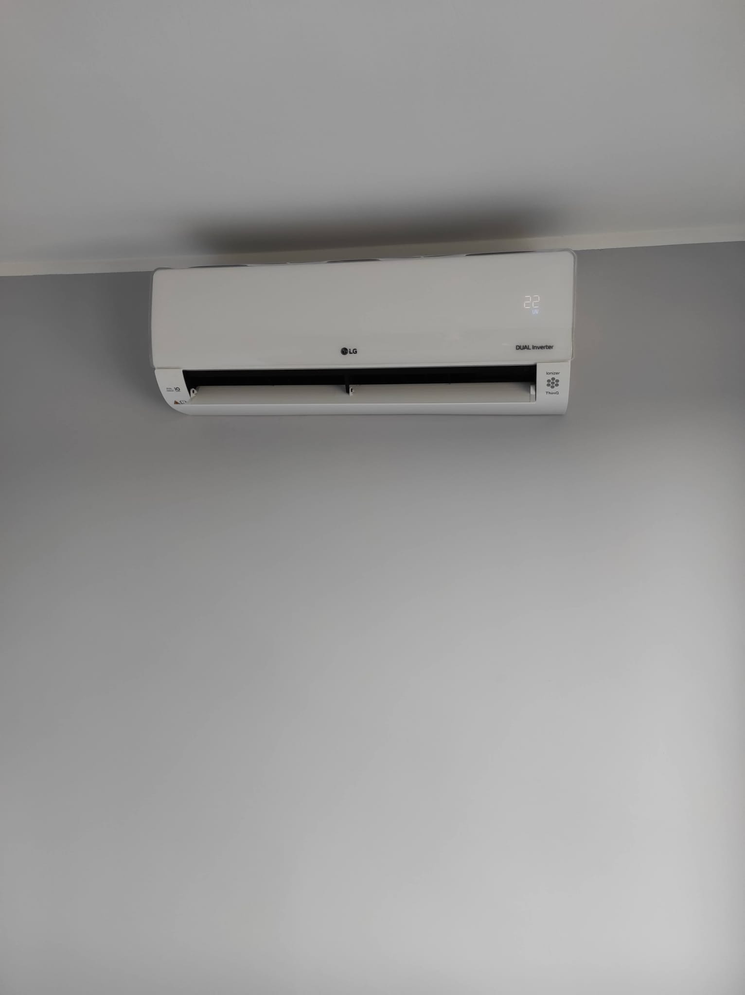 Biały klimatyzator LG Dual Inverter zamontowany na szarej ścianie, wyświetlający temperaturę 22 stopnie.