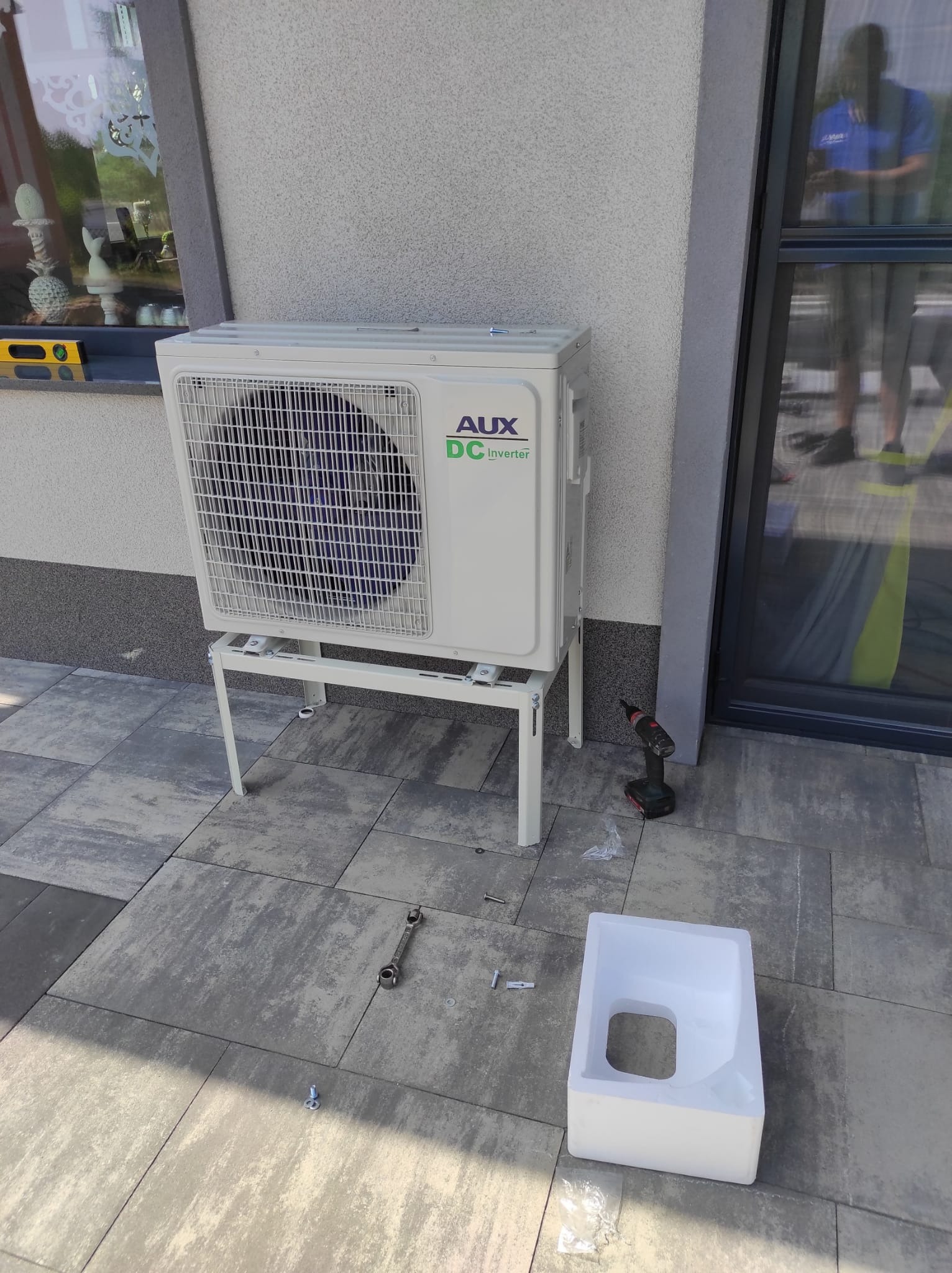 Biała jednostka zewnętrzna klimatyzatora AUX DC Inverter zamontowana na metalowym stelażu przed budynkiem, obok wkrętarka, klucz płaski i styropianowe opakowanie.