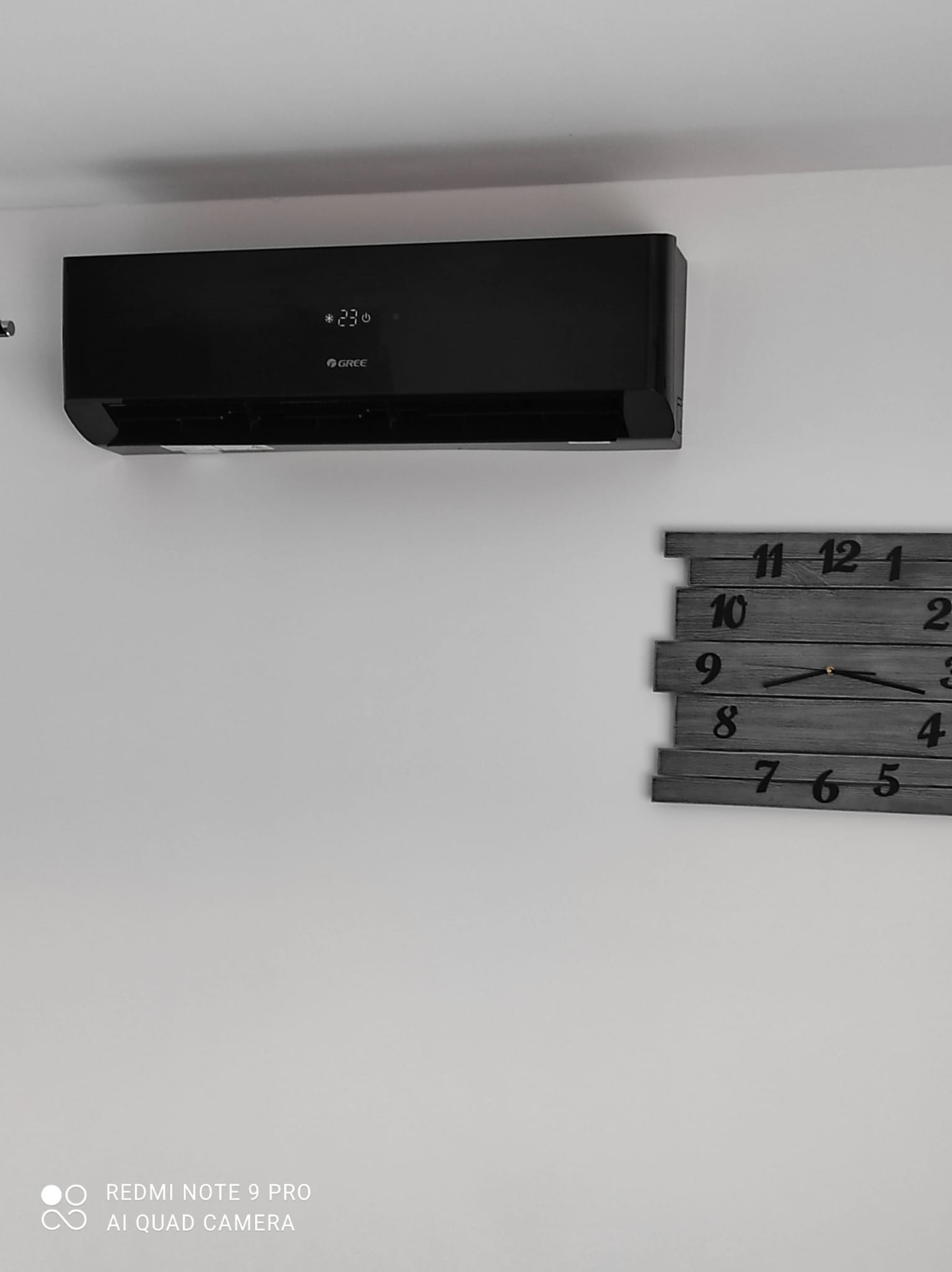 Czarny klimatyzator marki Gree zamontowany na białej ścianie, obok drewniany zegar z czarnymi cyframi.