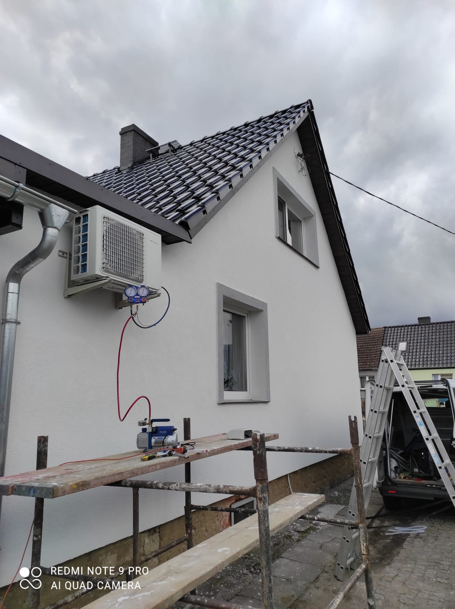 Biała jednostka zewnętrzna klimatyzacji zamontowana na ścianie domu, podłączona do manometrów i pompy próżniowej na rusztowaniu, drabina oparta o samochód dostawczy w tle.
