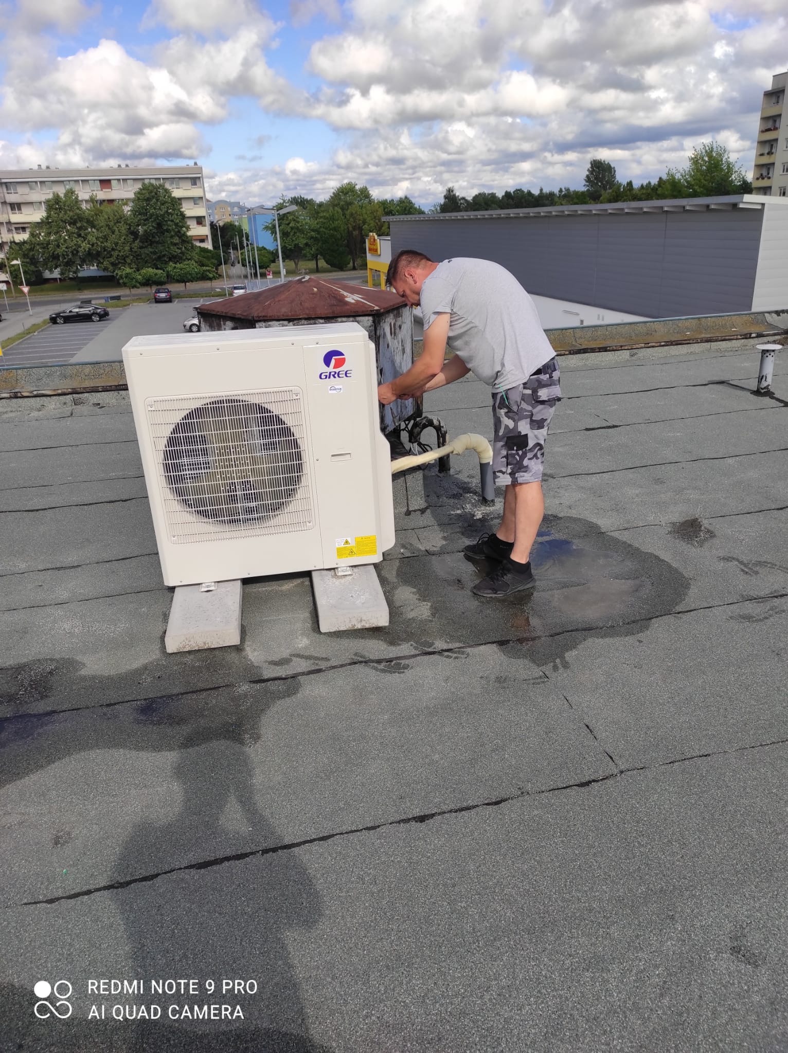 Mężczyzna w krótkich spodenkach w kamuflażu podłącza zewnętrzną jednostkę klimatyzacji marki Gree na płaskim dachu.