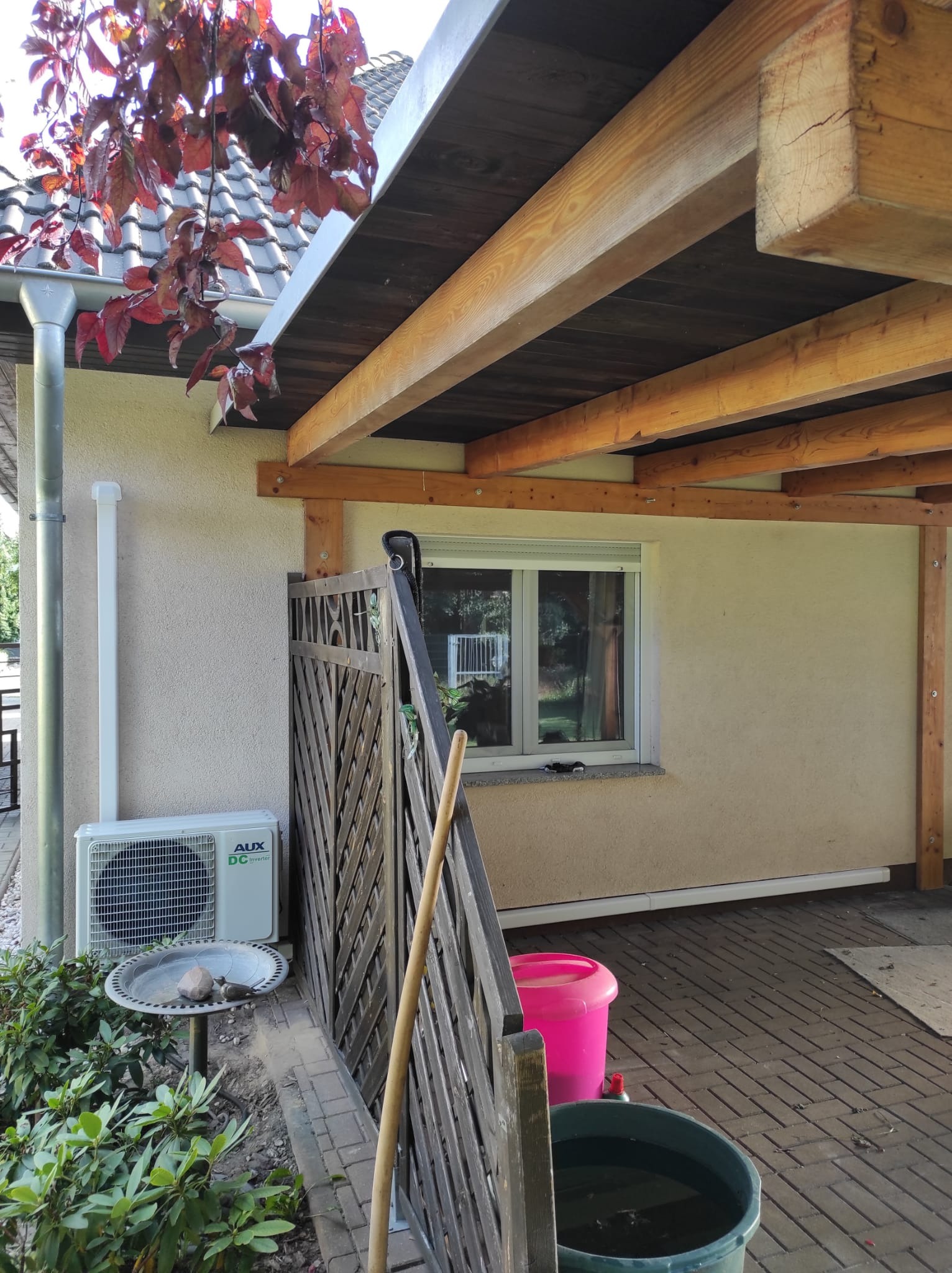 Zewnętrzna jednostka klimatyzacji AUX DC Inverter zamontowana przy ścianie domu w Gubinie, widoczna zza drewnianego płotu, obok poidełka dla ptaków i roślinności.