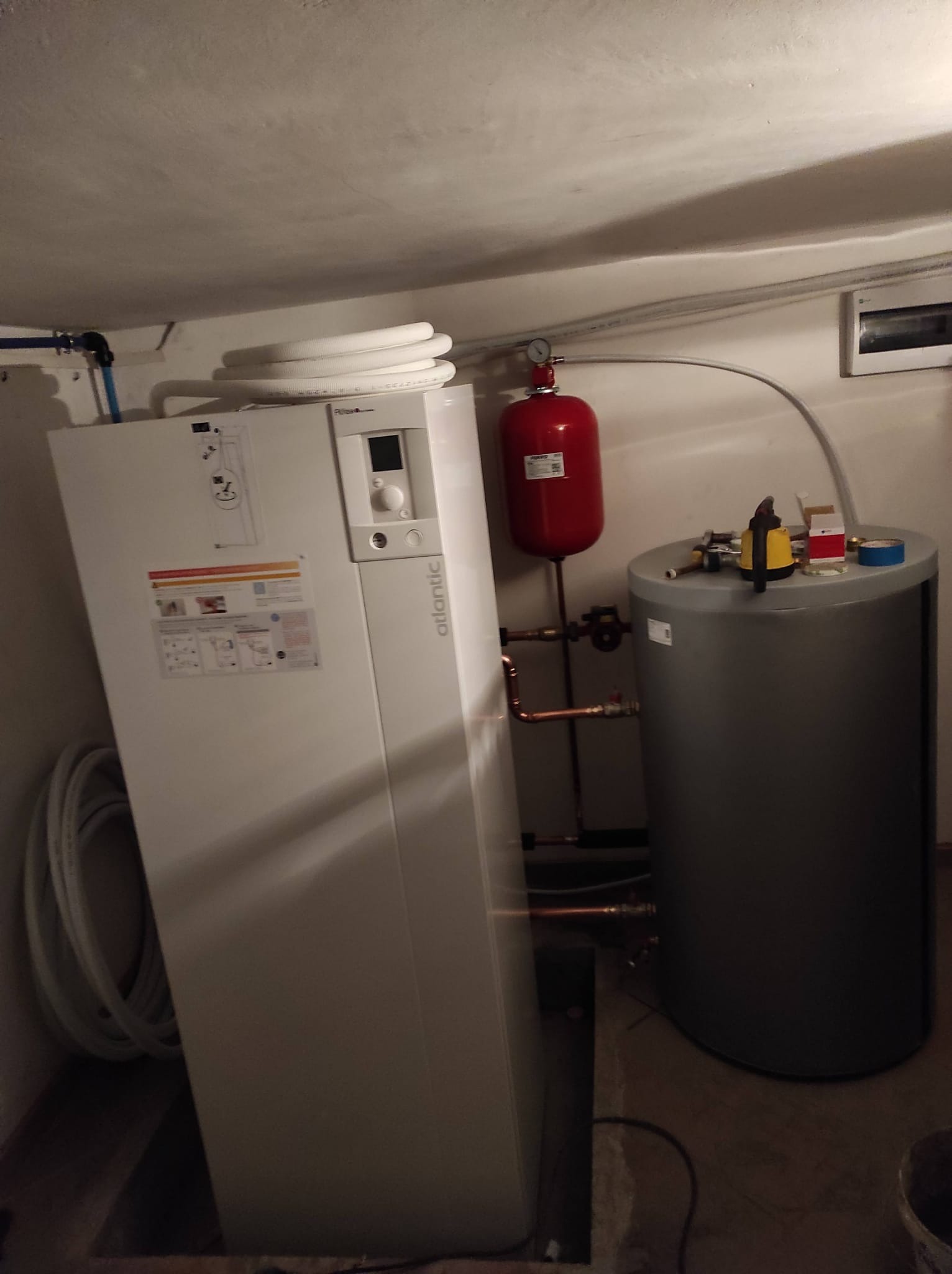 Biała pompa ciepła Atlantic z czerwonym zbiornikiem wyrównawczym i szarym zasobnikiem ciepłej wody w piwnicy, widoczne rury miedziane i narzędzia na zasobniku.