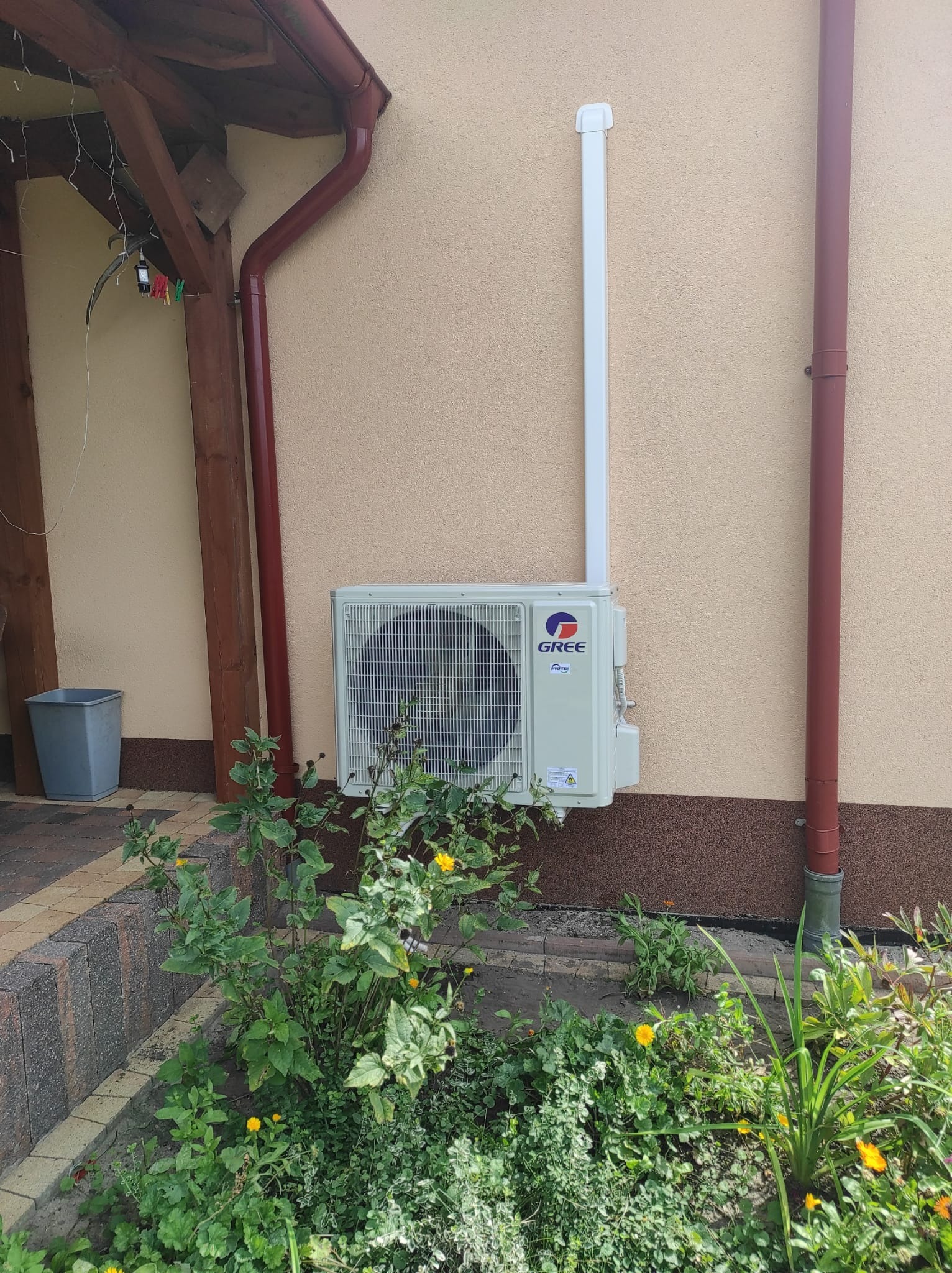 Zewnętrzna jednostka klimatyzacji marki Gree zamontowana na elewacji domu, widoczna z poziomu ogrodu z kwitnącymi roślinami, obok rynna spustowa w kolorze ceglastym.