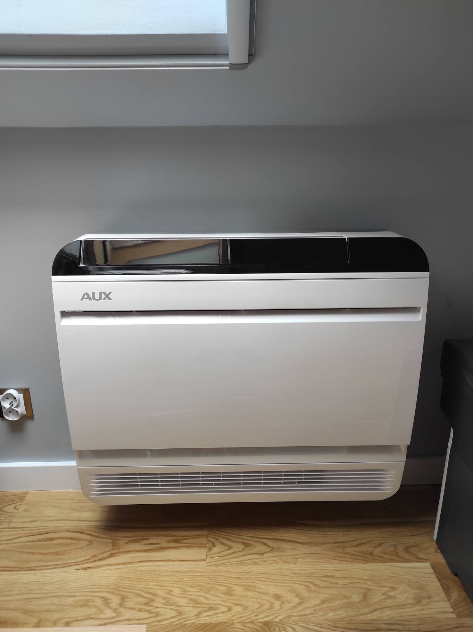Biała, stojąca jednostka klimatyzacji AUX zamontowana przy szarej ścianie, widoczne gniazdko elektryczne i drewniana podłoga.