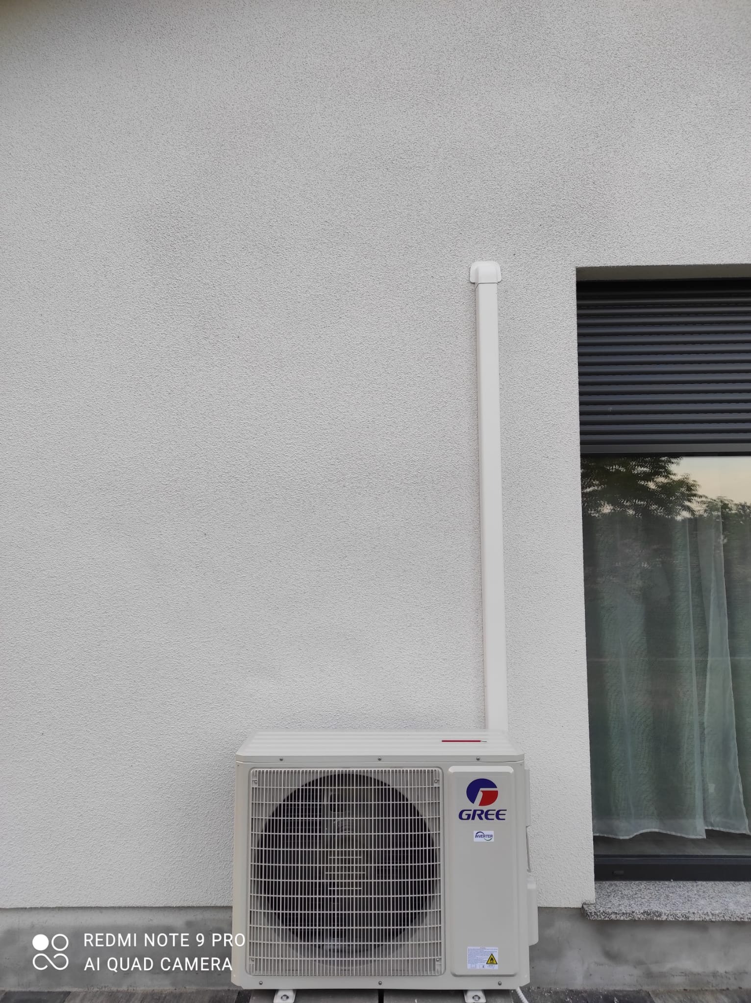 Zewnętrzna jednostka klimatyzacji marki Gree zamontowana przy ścianie budynku z oknem z żaluzjami zewnętrznymi.