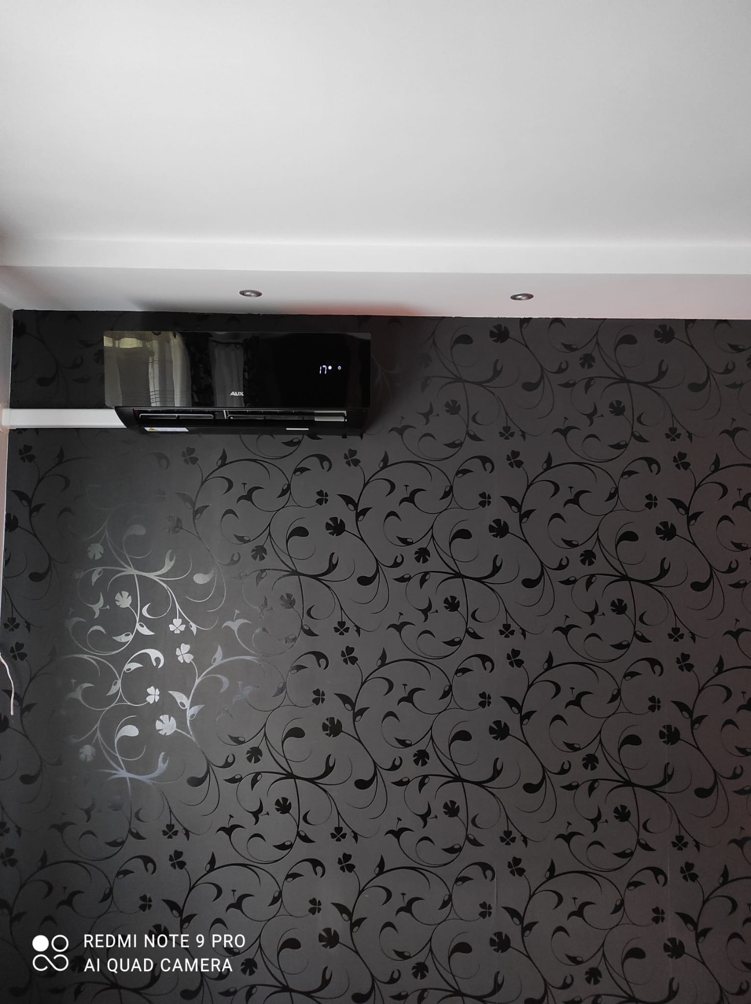 Czarny klimatyzator AUX zamontowany na ścianie pokrytej ciemną tapetą w kwiaty, wyświetlający temperaturę 17 stopni.
