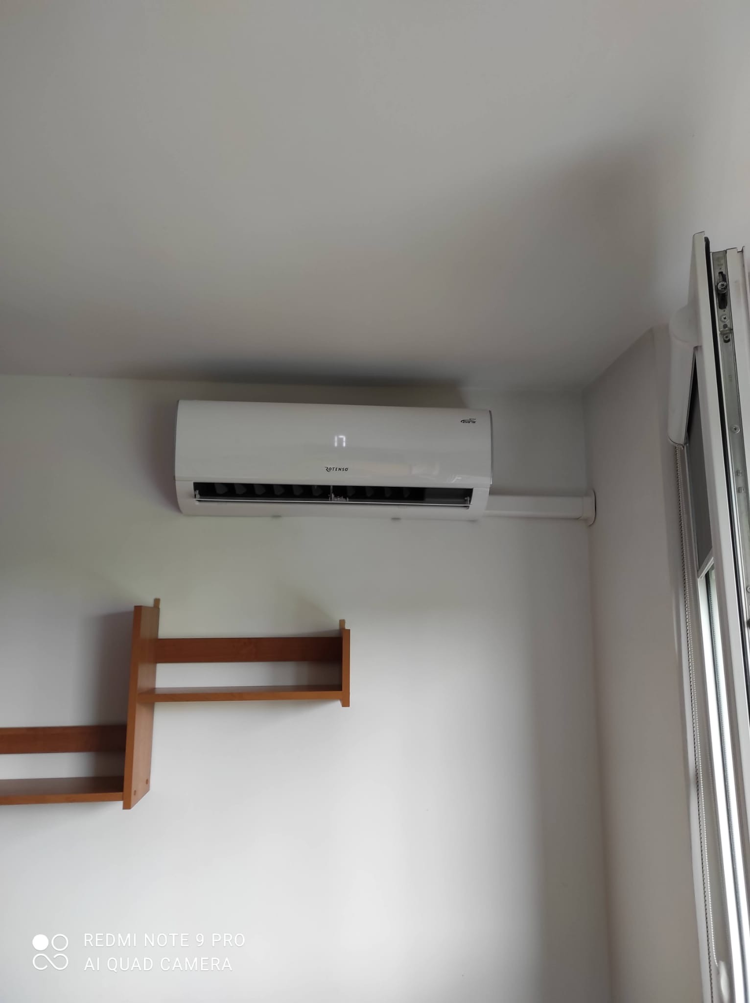 Biały klimatyzator Rotenso zamontowany na białej ścianie w pomieszczeniu, widoczna rurka odprowadzająca oraz półka z drewna.