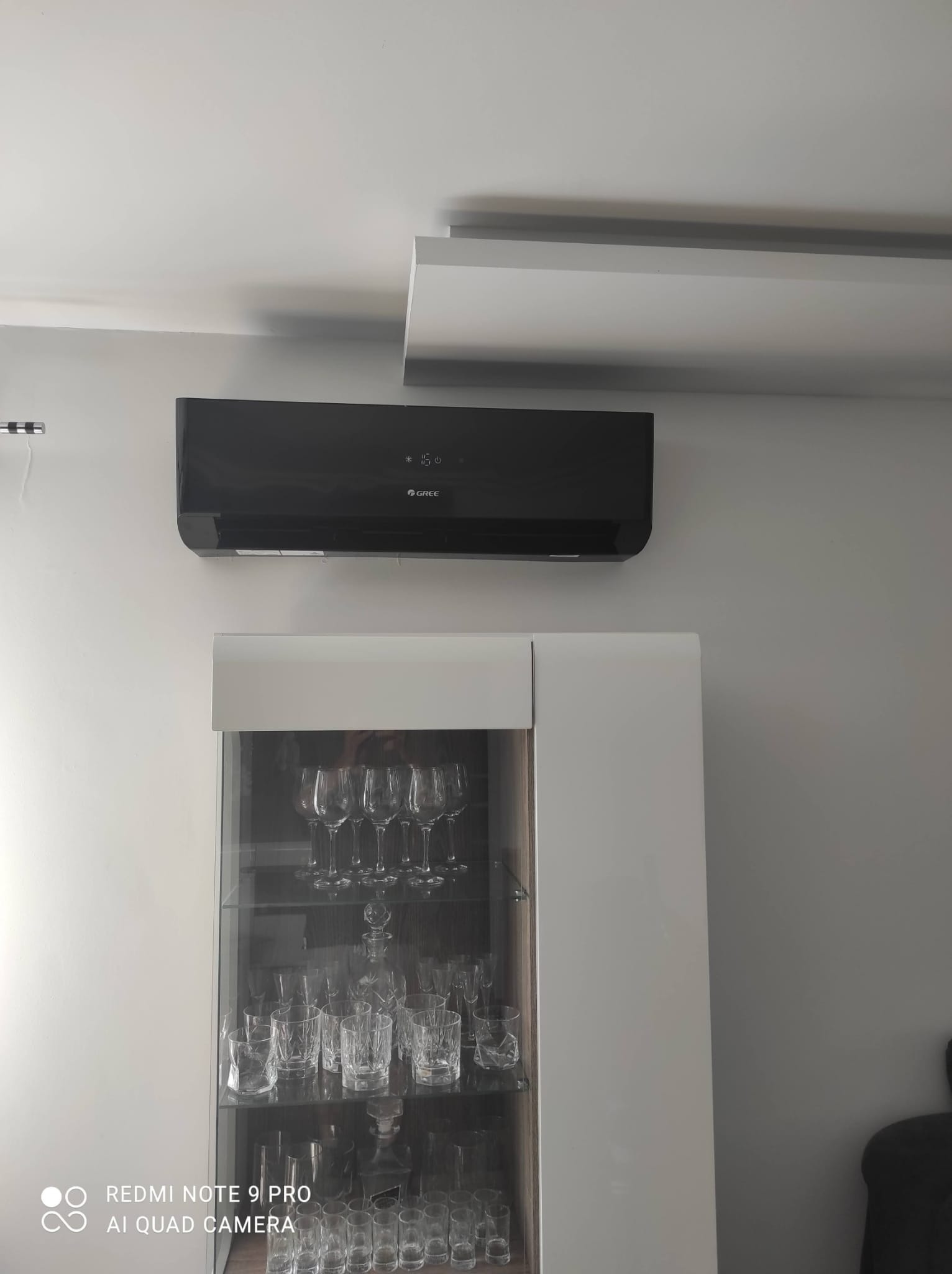 Czarny klimatyzator marki Gree zamontowany na szarej ścianie nad przeszkloną, białą witryną z kryształami i kieliszkami.