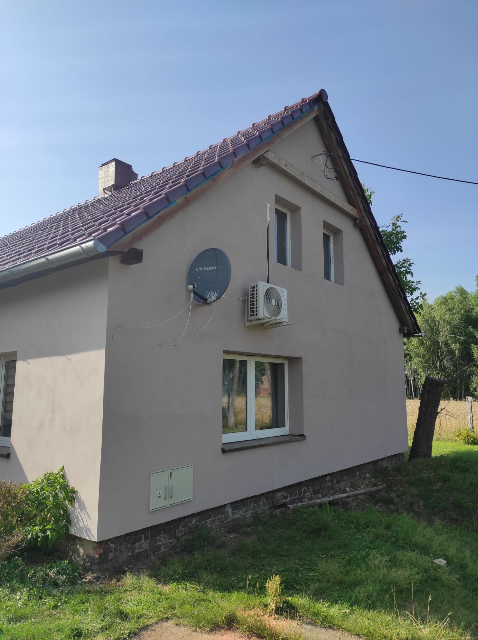 Szary dom jednorodzinny z zamontowaną jednostką zewnętrzną klimatyzacji na ścianie, obok okna na pierwszym piętrze i anteną satelitarną. Widoczny również fragment dachu z brązową dachówką i trawnik.