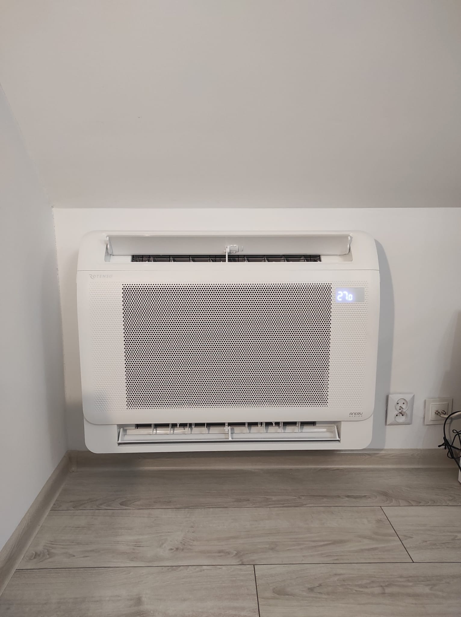 Biały klimatyzator ścienny marki Rotenso, model Aneri Series, z wyświetlaczem wskazującym temperaturę 27 stopni Celsjusza, zamontowany w pomieszczeniu z szarą podłogą imitującą drewno.