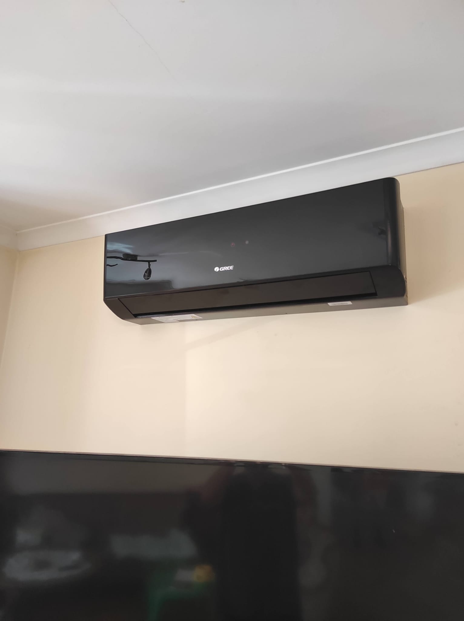 Czarna klimatyzacja marki Gree zamontowana na ścianie w jasnym pomieszczeniu, widoczny fragment telewizora pod spodem.