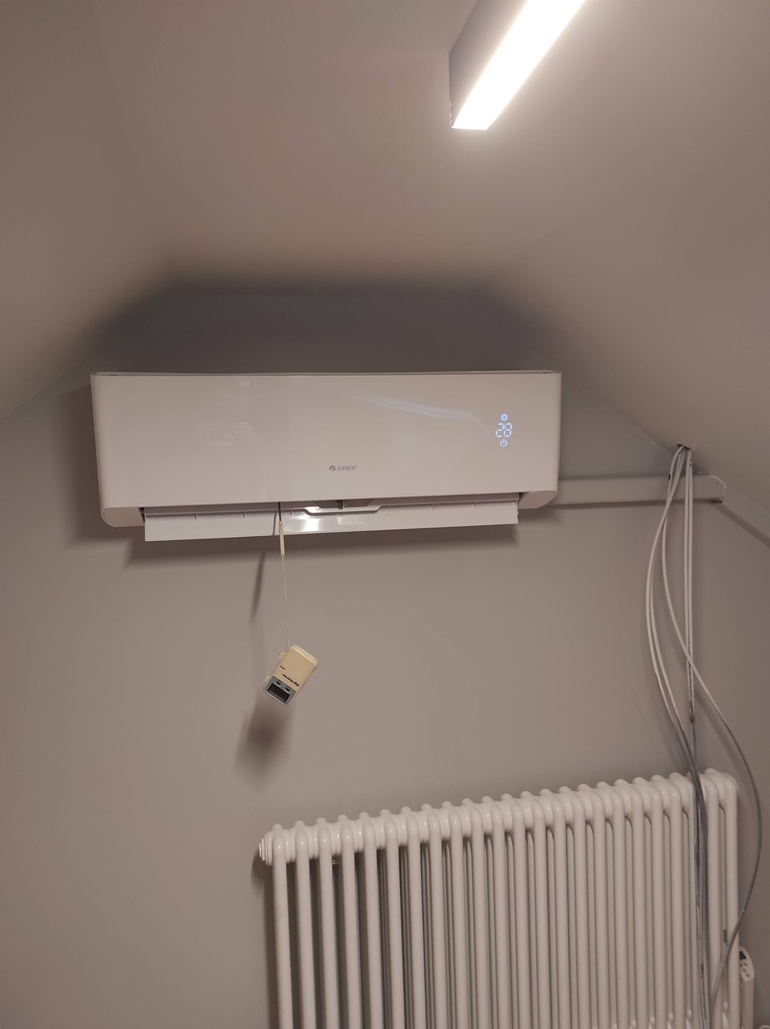 Biały klimatyzator marki Gree zamontowany na szarej ścianie pod skosem, widoczny biały grzejnik żeberkowy i zwisający sterownik na kablu.
