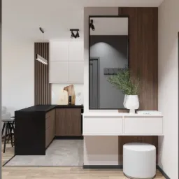 Wnętrze mieszkania z widoczną kuchnią z czarnym blatem i drewnianymi szafkami, lustrem w czarnej ramie, białymi szafkami i pufą, oraz szarą ścianą z drzwiami.