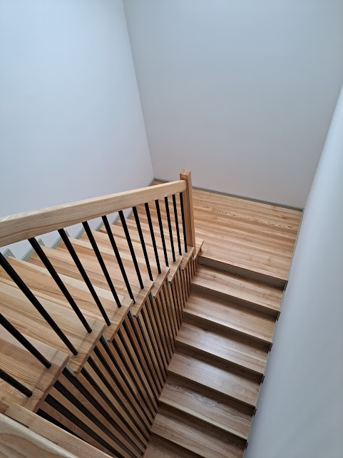 Jasne drewniane schody z czarnymi balustradami, widok z góry, minimalistyczne wnętrze z białymi ścianami.