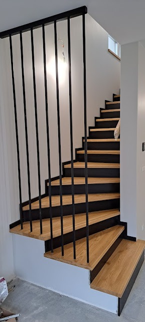 Nowoczesne schody z drewnianymi stopniami i czarnymi podstopnicami oraz metalową balustradą z pionowych prętów w jasnym wnętrzu, widoczne niedokończone wykończenie ścian.