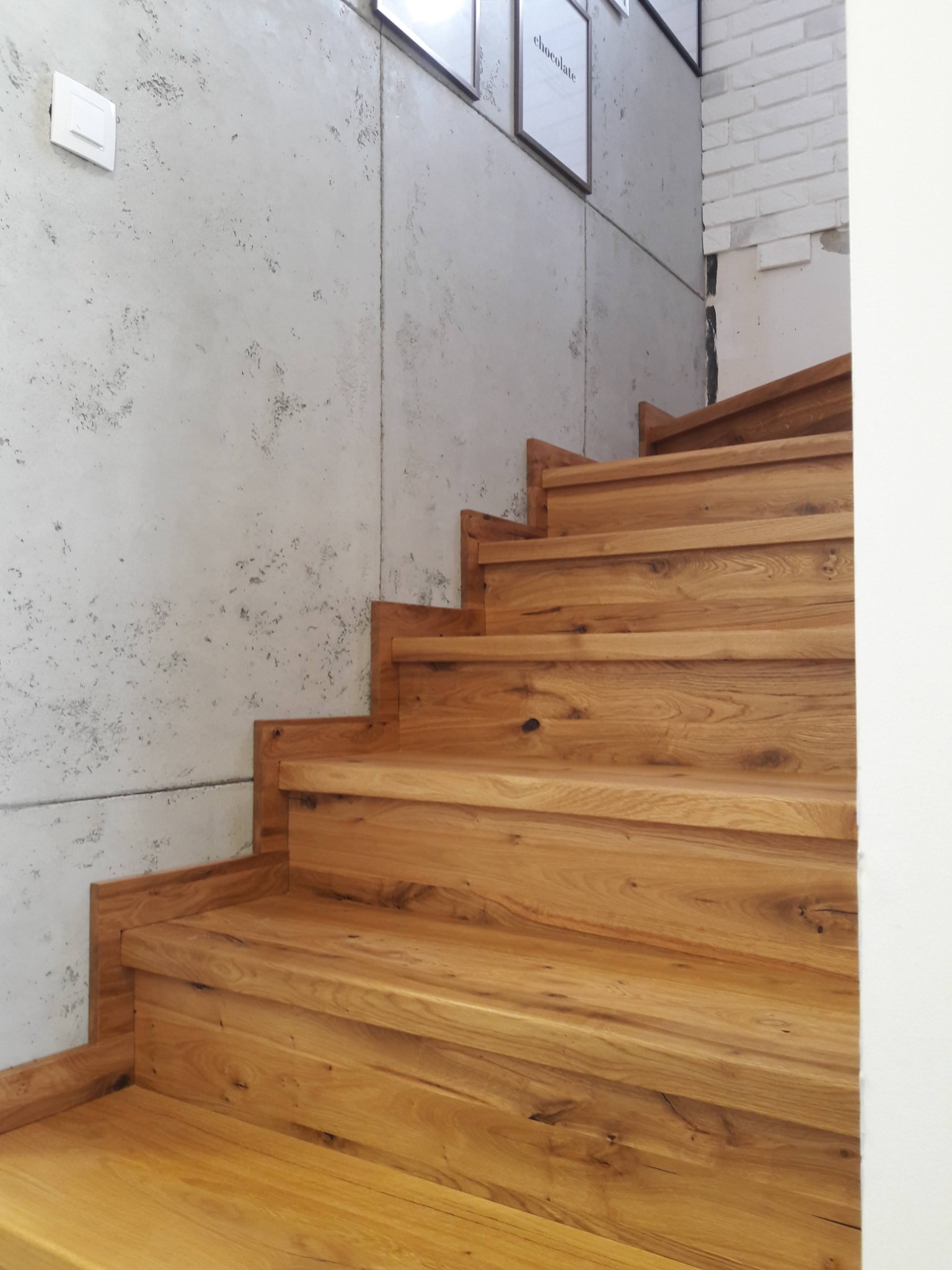 Jasne, drewniane schody z widocznym usłojeniem na tle betonowej ściany z włącznikiem i fragmentem ściany z białej cegły.