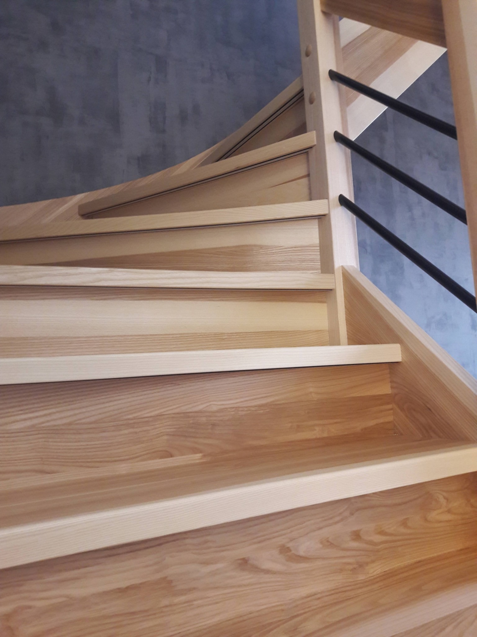 Jasne, drewniane schody z czarnymi, metalowymi balustradami na tle szarej ściany.