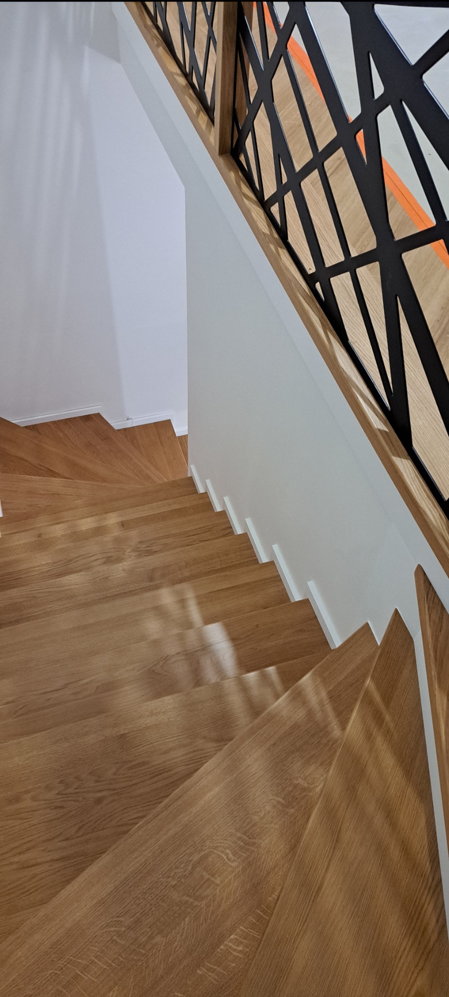 Drewniane schody z nowoczesną balustradą z czarnymi, geometrycznymi elementami, widok z góry na stopnie o ciepłym odcieniu drewna i białe ściany.