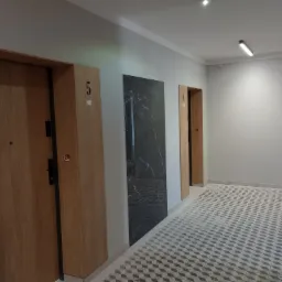 Korytarz w nowym budownictwie z trzema drzwiami o numerach 4 i 5, wykończony płytkami podłogowymi w geometryczny wzór oraz z czarną płytą dekoracyjną na ścianie.