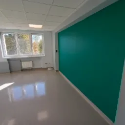Pomalowane na turkusowo ściany w pustym, jasnym pomieszczeniu biurowym z oknem i grzejnikiem, widoczne gniazdka elektryczne.