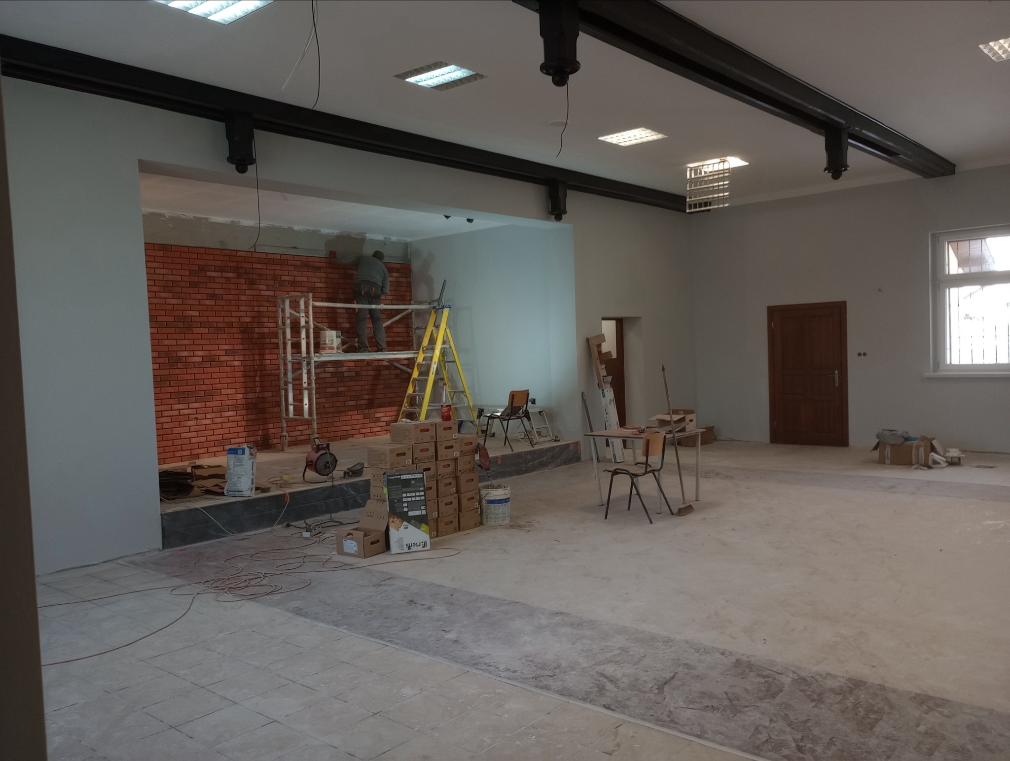 Mężczyzna na rusztowaniu układa cegiełki na ścianie w przestronnym, jasnym pomieszczeniu, widoczne skrzynki z materiałami, drabina i narzędzia.