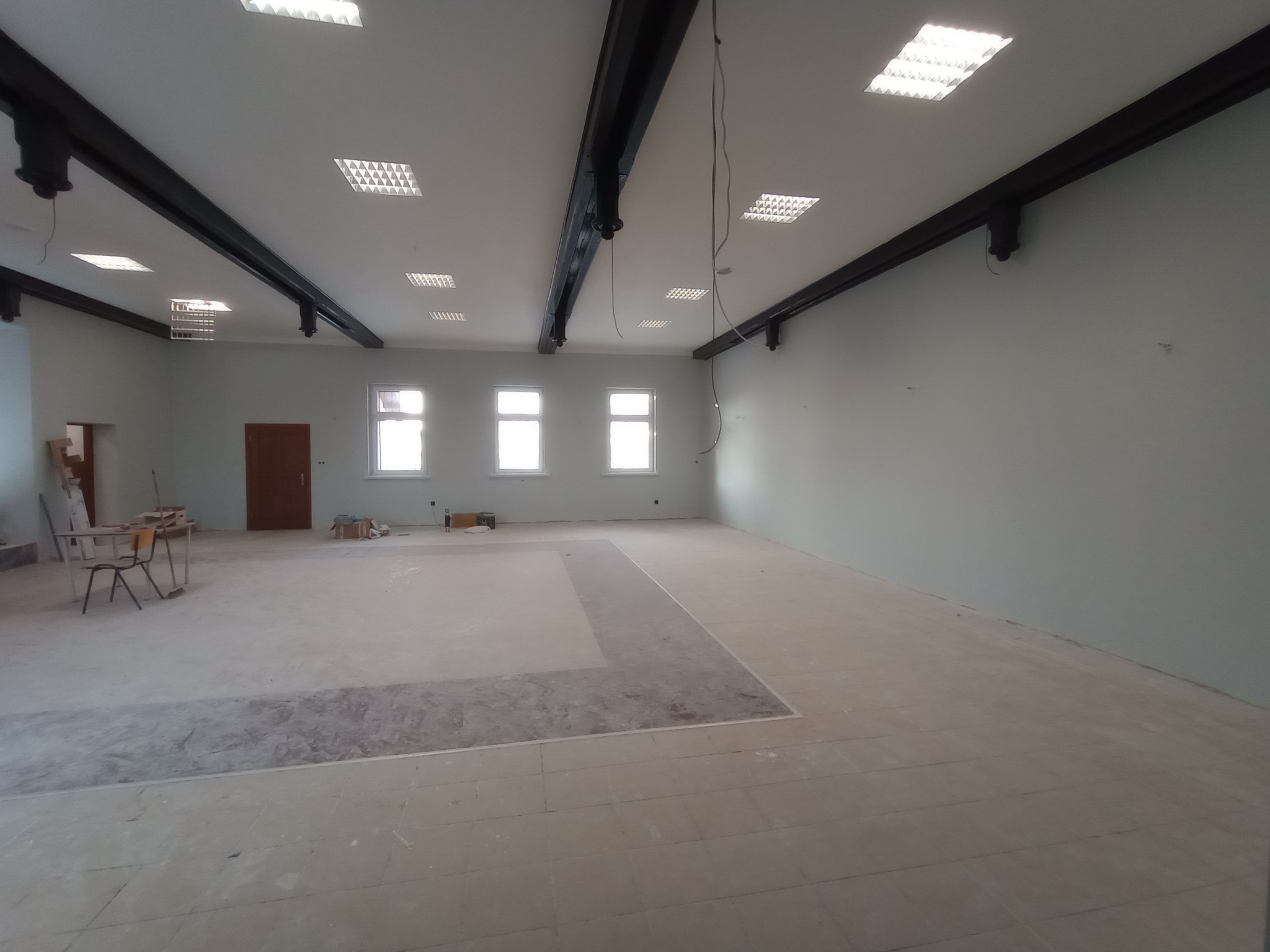 Duże, puste pomieszczenie w trakcie remontu z widocznymi fragmentami starej podłogi, przygotowane do dalszych prac wykończeniowych, oświetlone przez liczne lampy sufitowe.