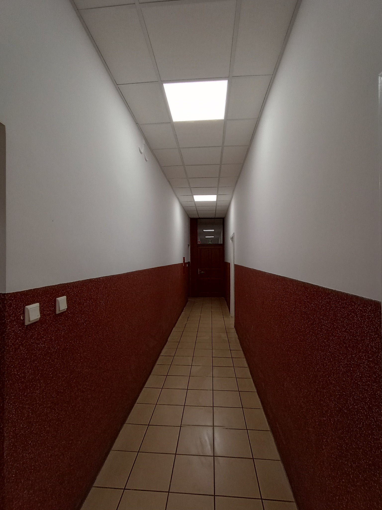 Długi korytarz biurowy z brązowymi ścianami do połowy wysokości, jasnymi płytkami podłogowymi i kasetonowym sufitem z wbudowanym oświetleniem, widok perspektywiczny.